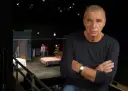 Dawid Głowacki, reżyser teatralny, z rękami skrzyżowanymi na piersi, na tle sceny z łóżkiem i ludźmi.