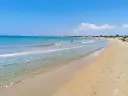 A wide, sandy beach with clear turquoise water and a few people swimming and sunbathing under a bright blue sky.