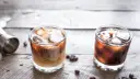 Dwa drinki na drewnianym stole: jeden to kremowy White Russian, drugi to ciemny Black Russian.