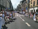 Procesja dzieci w białych strojach, niosących koszyki, idzie ulicą. W tle widać budynki i ludzi, co może symbolizować sekularyzację społeczeństwa.