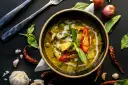 Zielone curry w misce, ozdobione chili i bazylią, otoczone przyprawami.