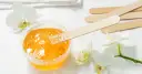 Słoneczna pasta cukrowa do depilacji w słoiczku, drewniane szpatułki i białe orchidee tworzą delikatny zestaw do pielęgnacji.