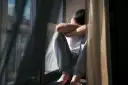 Mężczyzna siedzi przy oknie, obejmując się ramionami.