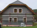 Dom w trakcie ocieplania. Szara elewacja, drewniane okna, rusztowanie. Materiały budowlane Austrotherm wskazują na dbałość o niski współczynnik przewodzenia ciepła.