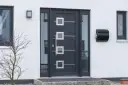 Nowoczesne drzwi zewnętrzne w ciemnoszarym kolorze z geometrycznymi przeszkleniami i metalowymi elementami. Obok skrzynka na listy.