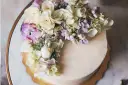 Elegancki tort z kremową polewą, ozdobiony bukietem żywych goździków, róż i hortensji w pastelowych kolorach.