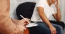 Dłoń pisząca na notatniku, w tle mężczyzna w białej koszulce.