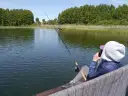 Osoba siedzi na pomoście i łowi ryby na jeziorze Jagodne. W tle las i trzciny.