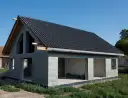 Dom w stanie surowym otwartym, z ciemnym dachem i szarymi ścianami z bloczków. Widać otwory okienne i drzwiowe, a także konstrukcję dachu.