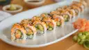 Rząd roladek sushi california roll, posypanych sezamem i ikrą, z awokado i łososiem w środku. Obok imbir marynowany i ogórek.