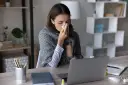 Chora kobieta z chusteczką przy nosie, otulona kocem, pracuje przy laptopie. Dyskutuje o swoim prawie do zasiłku chorobowego.