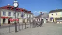Słoneczny stary rynek łowicz z brukowanym placem, budynkami z czerwonymi dachami i zegarem na słupie.