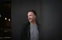 Joris, lächelnd, lehnt an einer dunklen Wand.