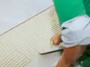 Ręka z kielnią rozprowadza klej na podłodze, tworząc wzory. To pokazuje, jaki grzebień do płytek jest używany do równomiernego rozprowadzenia zaprawy.