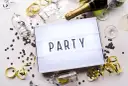Świętowanie zaczyna się od zaproszeń! Na stole leżą kieliszki, szampan i konfetti, a tablica świetlna głosi "PARTY".