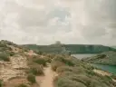 Malta czy warto? Kamienista ścieżka prowadzi przez skaliste wybrzeże z niską roślinnością do zatoki. W oddali widać wieżę obronną.