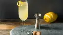 French 75 na ginie kontra French 75 na koniaku. Elegancki drink z cytrynową skórką, kieliszek, miarka barmańska, cytryna i korek od szampana.