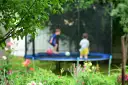 Dzieci bawią się na trampolinie w ogrodzie, otoczone zielenią i kwiatami.