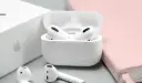 Białe słuchawki AirPods w etui, obok pudełka Apple i różowego notatnika.