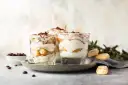 Pyszne desery włoskie: tiramisu w szklankach, udekorowane kakao i kawą. Obok kawałki biszkoptów i ziarna kawy.