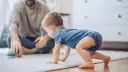 Dziecko w niebieskim body raczkuje na dywanie, obok siedzi ojciec z zabawkami.