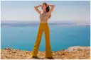 Kobieta w musztardowych spodniach dzwonach i beżowym topie na tle błękitnego jeziora.