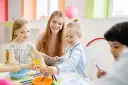 Uśmiechnięta opiekunka pomaga dzieciom malować. Warto wiedzieć, ile zarabia opiekun w żłobku, bo to praca pełna pasji.