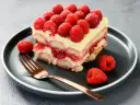 Pyszne tiramisu malinowe z malinami na wierzchu i na talerzu, obok złoty widelec.