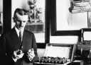 Nikola Tesla w garniturze trzymający dwa metalowe elementy, obok niego aparatura.