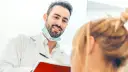 Uśmiechnięty dentysta w białym fartuchu i masce na szyi rozmawia z pacjentką, trzymając notatnik.