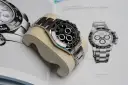 Luxuriöse Rolex Daytona Uhr auf einem Kissen vor einem Buch mit Uhrenabbildungen.