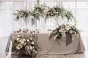 Eleganckie ścianki na wesele ozdobione bujnymi kwiatami i zielenią. Stół przykryty szarym obrusem, udekorowany świecami i kompozycjami kwiatowymi.