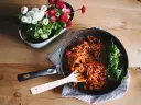 Patelnia ze spaghetti w sosie pomidorowym, obok rukola i doniczka z kwiatami.