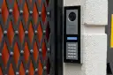 Domofon z kamerą i klawiaturą numeryczną zamontowany na ścianie obok czarnej kraty.