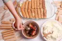 Dziecko zanurza biszkopty w kawie, przygotowując warstwy do tiramisu. Obok krem i kakao.