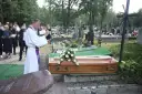 Wojciech Magnuski żegna zmarłego na cmentarzu. Ksiądz w białej szacie czyta z księgi przy trumnie ozdobionej białymi różami.