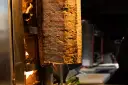 Duży pionowy obrotowy grill z mięsem, obok płomienie ognia.