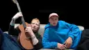 Ed Sheeran z gitarą i młody mężczyzna w niebieskiej bluzie siedzą na kanapie.