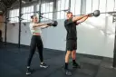 Kobieta i mężczyzna wykonują ćwiczenia z kettlebellami w siłowni.