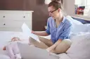 Kobieta w okularach siedzi na łóżku z laptopem i dokumentami, obok śpi dziecko. Załatwia formalności związane ze zmianą meldunku dziecka.