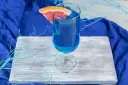 Przepis na Blue Lagoon: Błękitny drink z plasterkiem grejpfruta, ozdobiony niebieskimi gałązkami, na tle granatowej tkaniny.