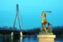 Warszawska Syrenka, bohaterka legendy, stoi dumnie z mieczem i tarczą. W tle widać nowoczesny most.