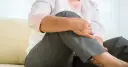 Kobieta w jasnej bluzce i szarych spodniach trzyma się za kolano.