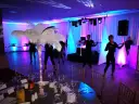 Impreza w stylu lat 20: tancerze, dekoracje z piór, stoły z kieliszkami i butelkami, kolorowe światła.