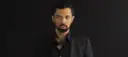 Mężczyzna w czarnym garniturze, z zarostem i kolczykiem w uchu. Jego spojrzenie sugeruje, że posiada najwyższy głos męski.