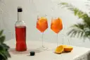 Aperol przepis: butelka Aperolu, dwa drinki z pomarańczą i lodem, połówki pomarańczy. Idealne na letnie popołudnie.