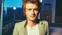 Joe Keery w jasnej marynarce, z naszyjnikiem, na tle nocnego miasta.