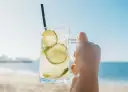 Orzeźwiająca Caipirinha czy Caipiroska z limonką i lodem w dłoni na tle plaży i morza. Idealny napój na upalny dzień.