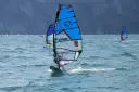 Osoba na desce windsurfingowej z niebieskim żaglem na wzburzonym morzu.