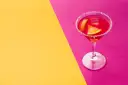 Elegancki cosmopolitan drink co zamiast żurawiny, z pomarańczą i lodem, na tle żółto-różowym.